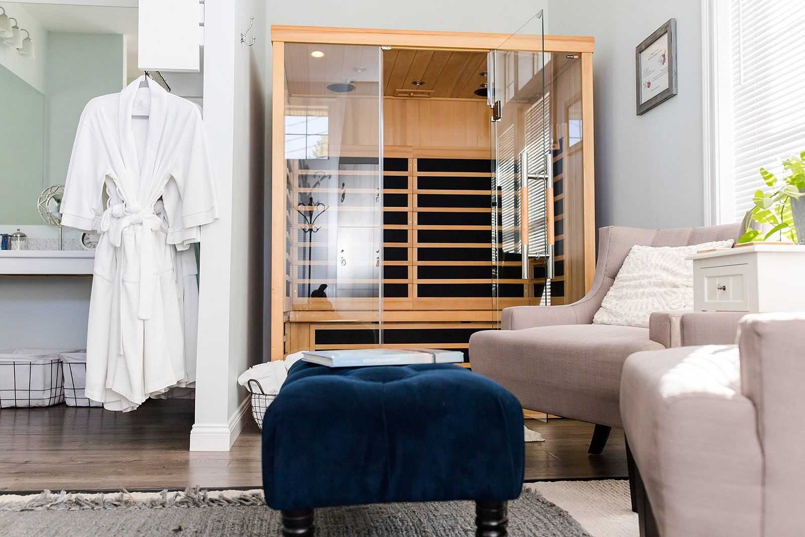 Modern spa room with a sauna, cozy seating, a bathrobe hanging, and a blue ottoman in the center.